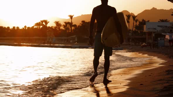 Silhouette Man Surfer in Sunset Light Walking Down Beach with Surfboard Near Waters Edge Enjoying