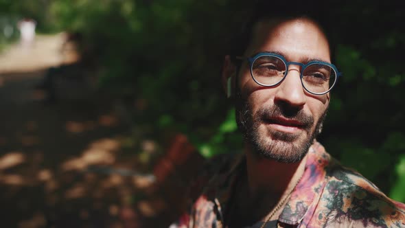 Relaxed curly-haired bearded man in eyeglasses listening music in headphones on the bench