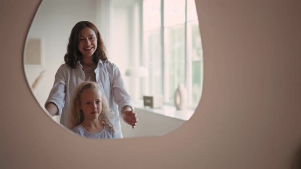 Beautiful Smiling Girl with Blond Curly Hair Looking at Her Reflection in Mirror While Her Mother