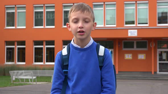 A Young Boy Talks To the Camera in Front of an Elementary School