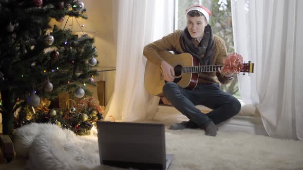 Wide Shot of Positive Young Man in Christmas Hat Waving at Laptop Video Chat and Playing Guitar