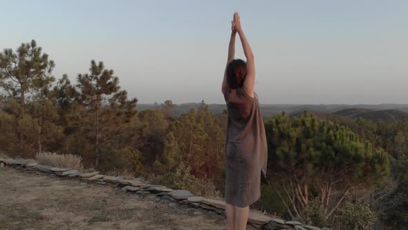 Woman practicing yoga in front of a cliff at the top of a hill with a scenic sunrise landscape durin