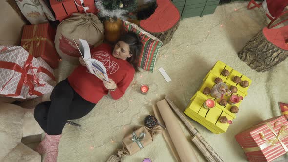 Pregnant Woman Reading a Book Under the Christmas Tree with Shining Christmas Lights