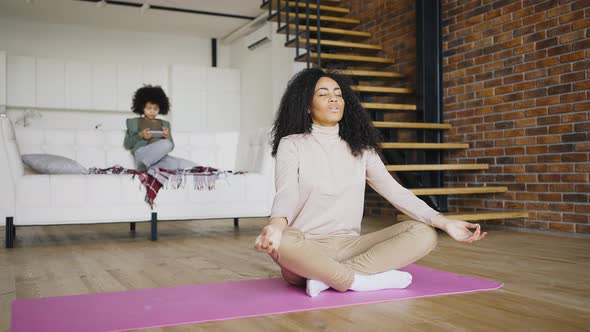Young Beautiful Curly Haired Woman Meditates While Her Son Plays with a Gadget
