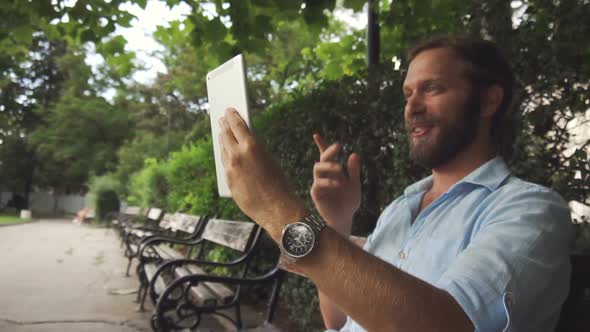 Nice looking man video chat on tablet on bench at park