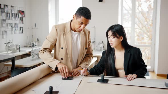 African American Man and Asian Woman Tailors Discussing Pants Pattern at Workshop Creating Fashion