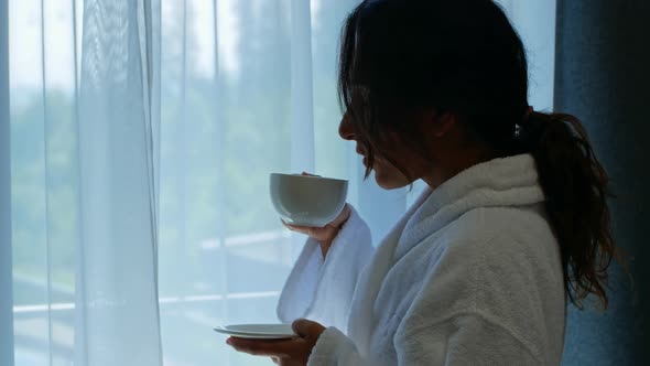 A beautiful young dark-skinned mixed-race woman stands by the window in the hotel and drinks coffee.