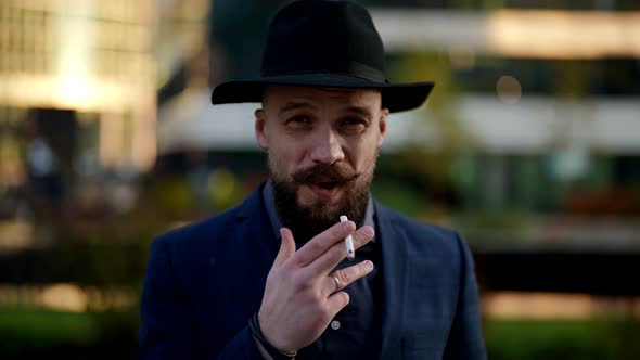 Portrait of a Bearded and Moustached Smoking Man in a Hat and a Plaid Jacket Against the Background