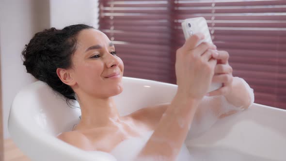 Young Happy Hispanic Woman Lying in Hot Comfortable Foam Luxurious Bath Relaxed Girl Typing Sms on