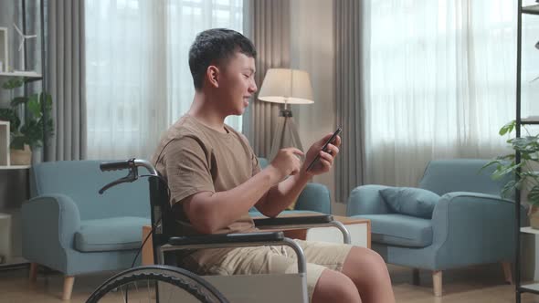 Young Asian Boy Sitting In A Wheelchair While Smiling And Using Smart Phone At Home alt