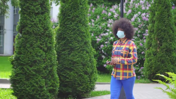 Afro American Woman with an Afro Hairstyle in a Protective Medical Mask with a Smartphone in His