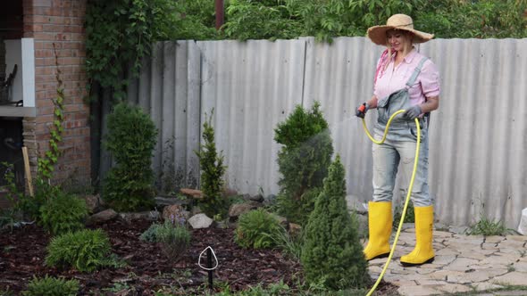 A Young Adult Woman Wateres a Vegetable Garden with a Garden Hose Outside the House