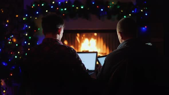 Father and Son Use Laptop and Tablet, Order Christmas Gifts While Sitting By the Fireplace