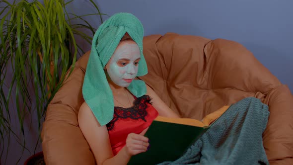 Woman with Towel on Head Mask on Face Sits and Reads Book in Armchair