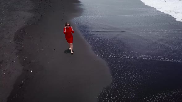 Aerial View of a Girl in a Red Dress Walking on the Beach with Black Sand