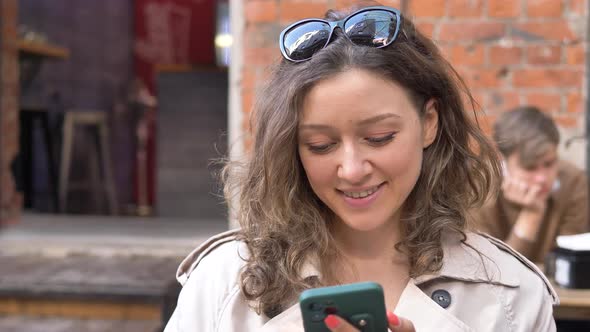 Young Woman Texts in Smartphone and Drinks Coffee in Cafe