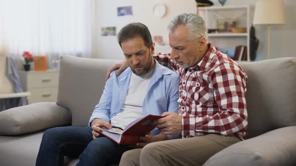 Pensive Adult Males Holding Album and Viewing Photos Recalling Relatives