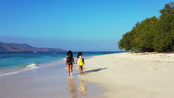 Two girls walking barefoot on wet sand of shore washed by white waves coming from blue turquoise sea