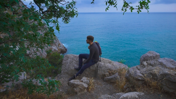 Young Hipster Guy Sitting On The Rock Mountains Near Sea