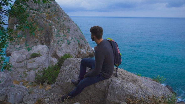 Young Hipster Guy Sitting On The Rock Mountains Near Sea, Man With a Rucksack On His Back