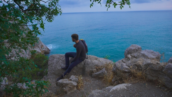 Young Hipster Guy Sitting On The Rock Mountains Near Sea, Man With a Rucksack On His Back