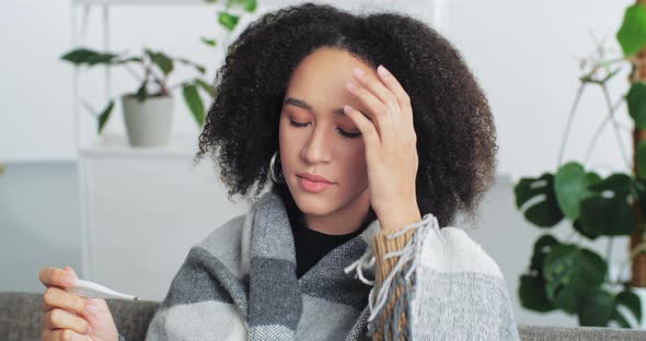 Young Sad Afro American Woman Feels Bad and Measures Her Temperature Sitting on Couch Sofa at Home