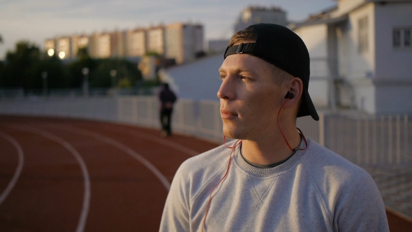 Young Man Wears Headphones On Field At Sunset On Stadium Track
