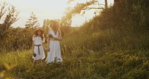 Two Girl Teenagers in White Dresses and Straw Hats with Flowers Bouquet Posing on Nature Landscape