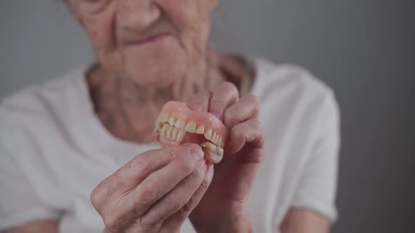 Toothless Senior Woman Holds Denture in Her Hands and Makes a Funny Chewing Movement with Her Teeth