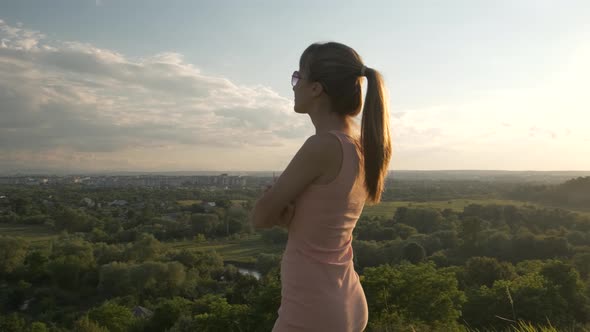 Young Woman in Pink Summer Dress Standing in Green Meadow Enjoying Sunset View