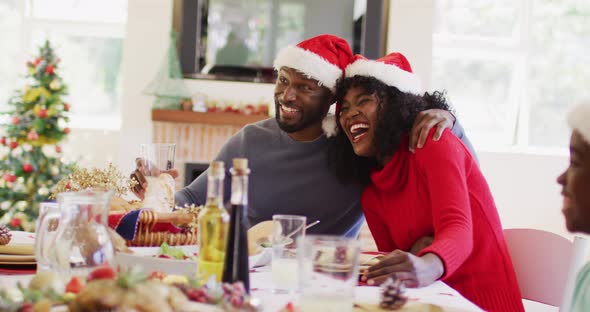 African american couple in santa hats smiling and toasting while sitting on dining table having lunc