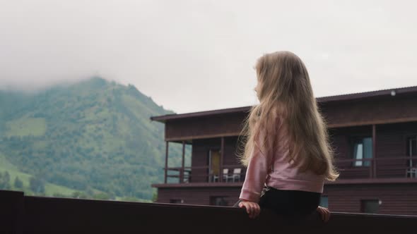 Cute Little Girl Sits on Railing Against Distant Mountains