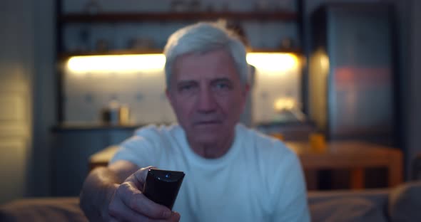 Close Up of Senior Man Using Tv Remote Control with Wife Cooking on Background