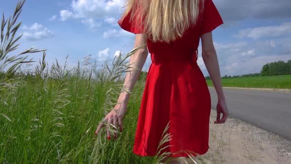 Young Beautiful Girl in a Red Dress Walks Along the Side of the Road and Touches the Green Plants