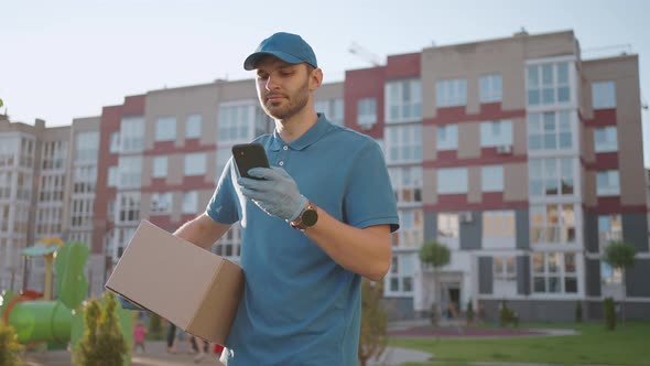 The Postman with Glasses Carries the Parcel and Looks at the Delivery Address Via Mobile Phone