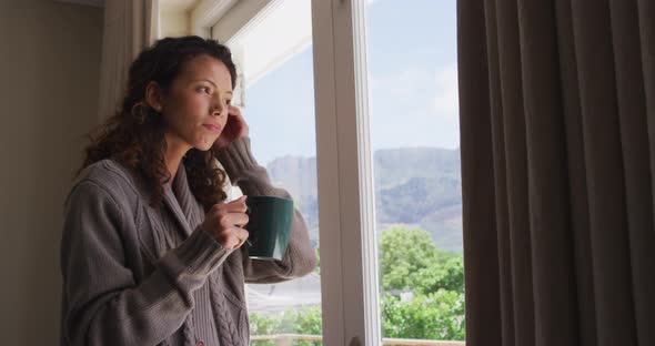 Thoughtful biracial woman standing with coffee at window and looking into distance