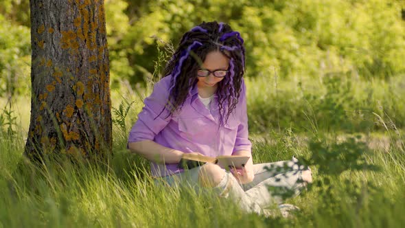 A woman in a purple shirt and glasses reads a literature under a tree.