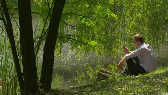 Man With Mobile Phone Sits to a Ground Lake Bank