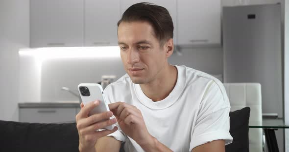 Close Up of Cheerful Young Man Sitting on the Sofa Using Smartphone Smile in the Modern Apartment