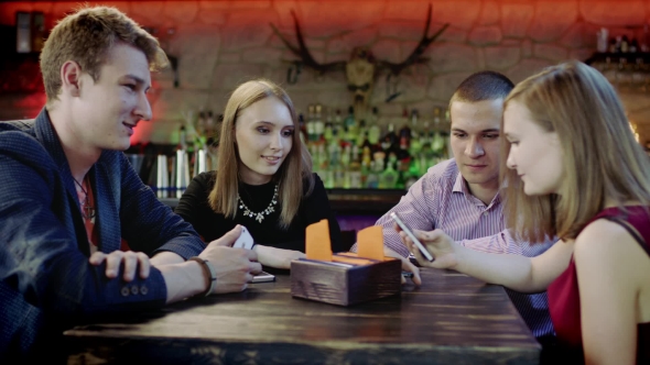 Company Of Young People Is Watching Something In The Cellphone Sitting In The Bar