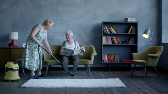 Elderly Woman Vacuuming The Floor, And An Elderly Man Reading a Newspaper