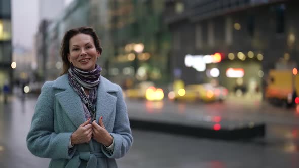 Portrait of a Smiling Woman in a Light Coat and Scarf at Dusk Against the Background of a Blurred