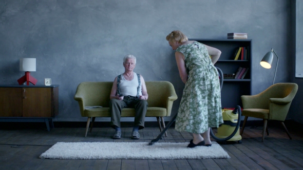 Elderly Woman Vacuuming The Floor, And An Elderly Man