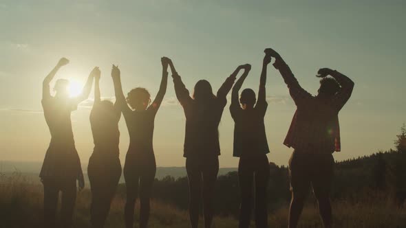 Silhouette of Diverse Multiracial Friends Standing with Arms Raised on Mountain Peak at Sunset