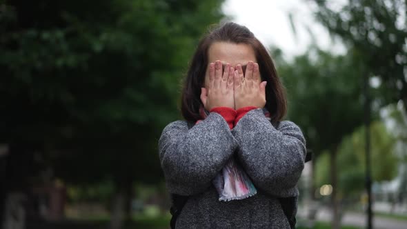 Young Caucasian Little Woman Closing Face with Hands Making Wish Standing Outdoors on Overcast Rainy