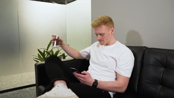 young caucasian male in white t-shirt reading his phone, man drinking water