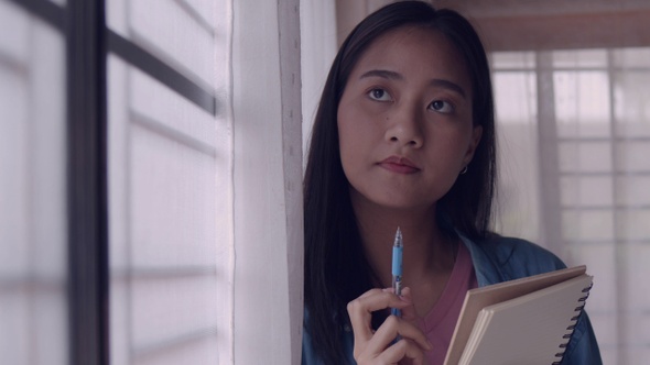 Young Asian businesswoman standing beside the window and writing in a notebook at the home office.