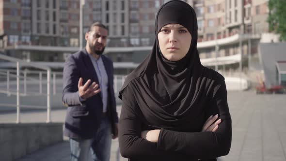 Portrait of Sad Woman in Hijab Standing with Hands Crossed and Looking at Camera As Angry Man