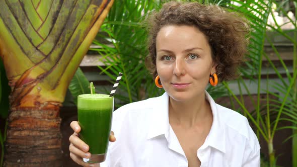 A Young Caucasian Woman Holds a Organic Cold Pressed Green Juice and Looking to the Camera