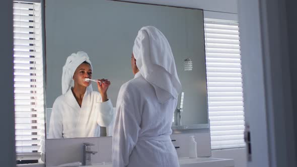 African american woman in bathrobe brushing her teeth looking in the mirror at bathroom